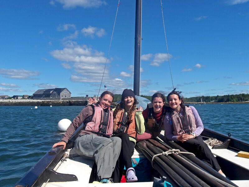 Four young people are on a sailboat, smiling at the camera. They are wearing life jackets, and the boat is on the water with a clear blue sky and some clouds in the background. The boat has a mast and rigging, and there are some buildings visible on the shore in the distance.